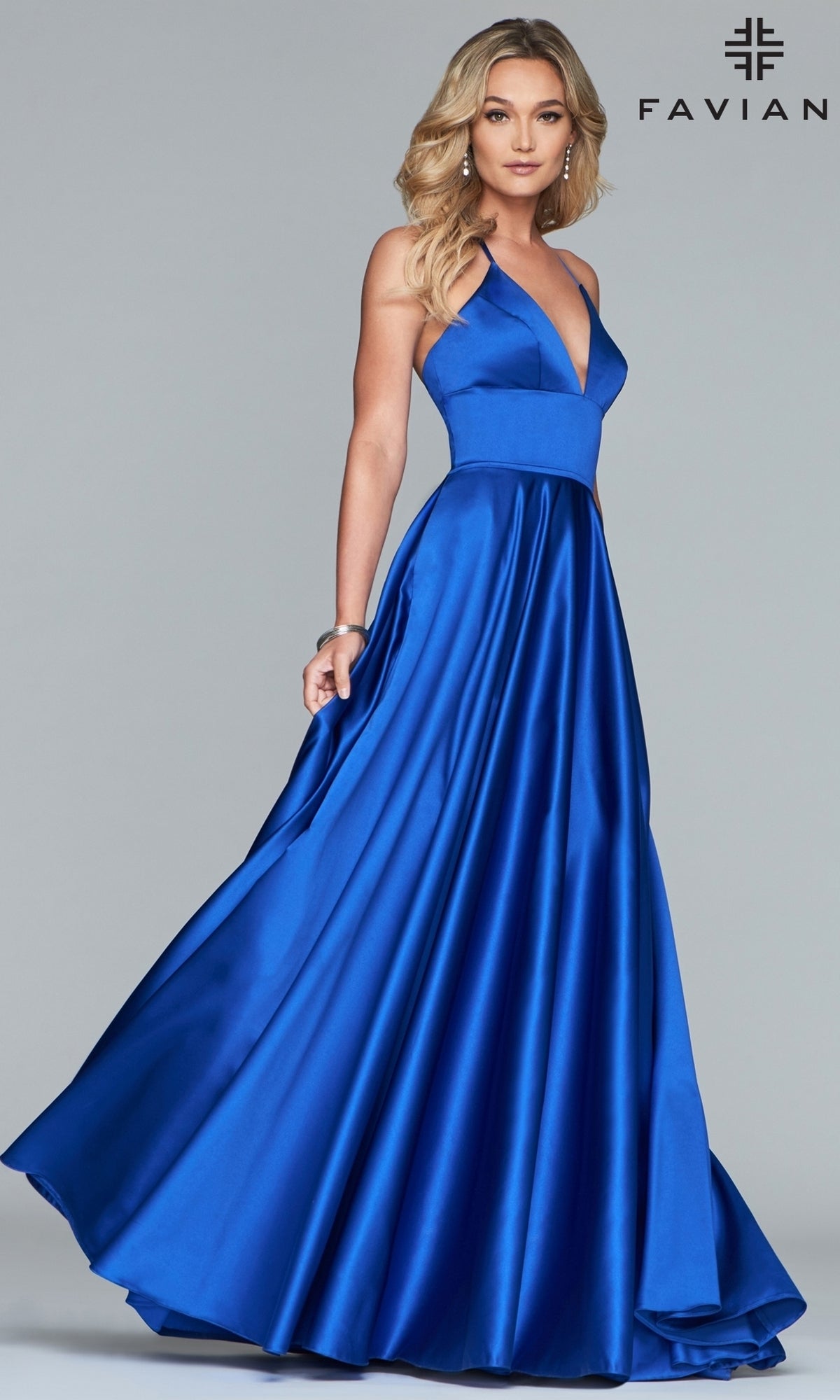 A woman poses against a gray background wearing the Corset-Back A-Line Long Satin Prom Dress by Faviana, featuring a sleeveless deep V-neck royal blue design. The Faviana logo appears in the top right corner.